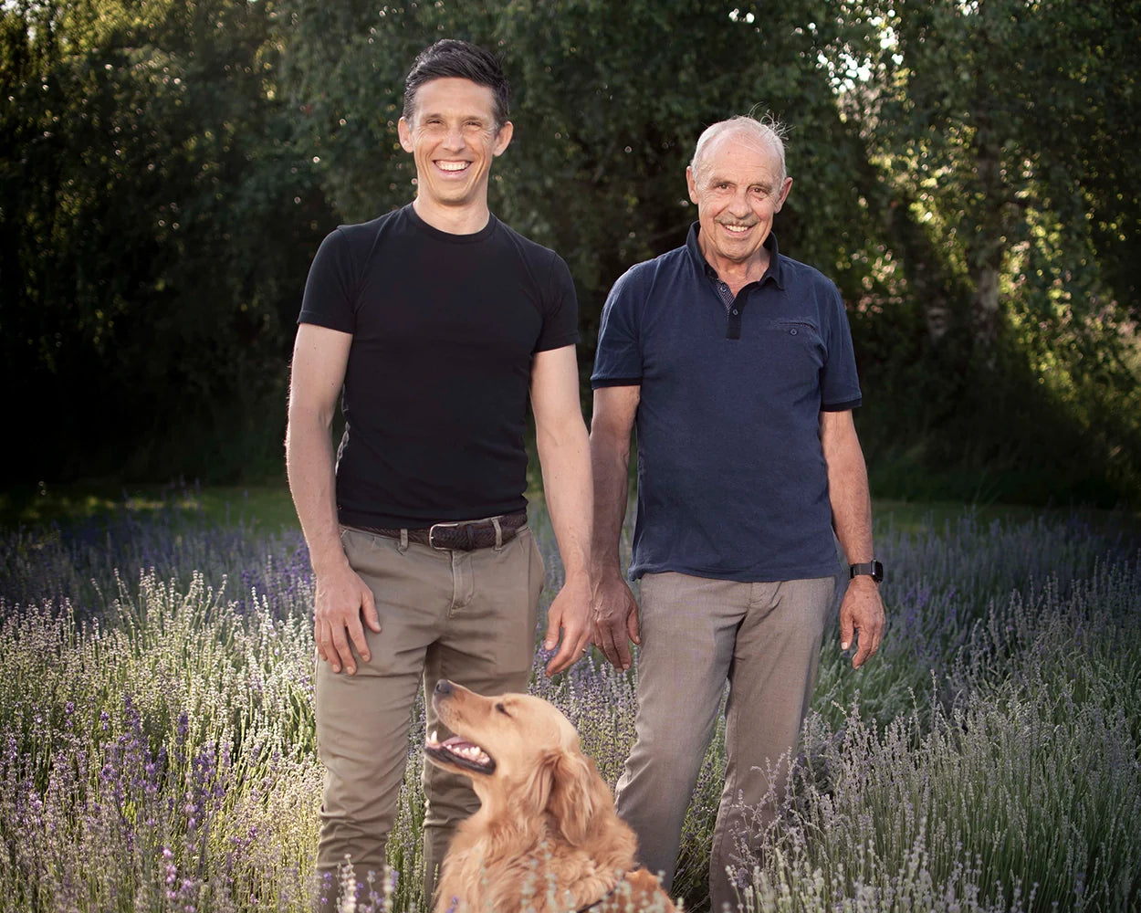 Zwei Männer stehen lachend in blühendem Lavendelfeld, Hund blickt aufmerksam, Galerie Position 1, Schweizer Aromatherapie