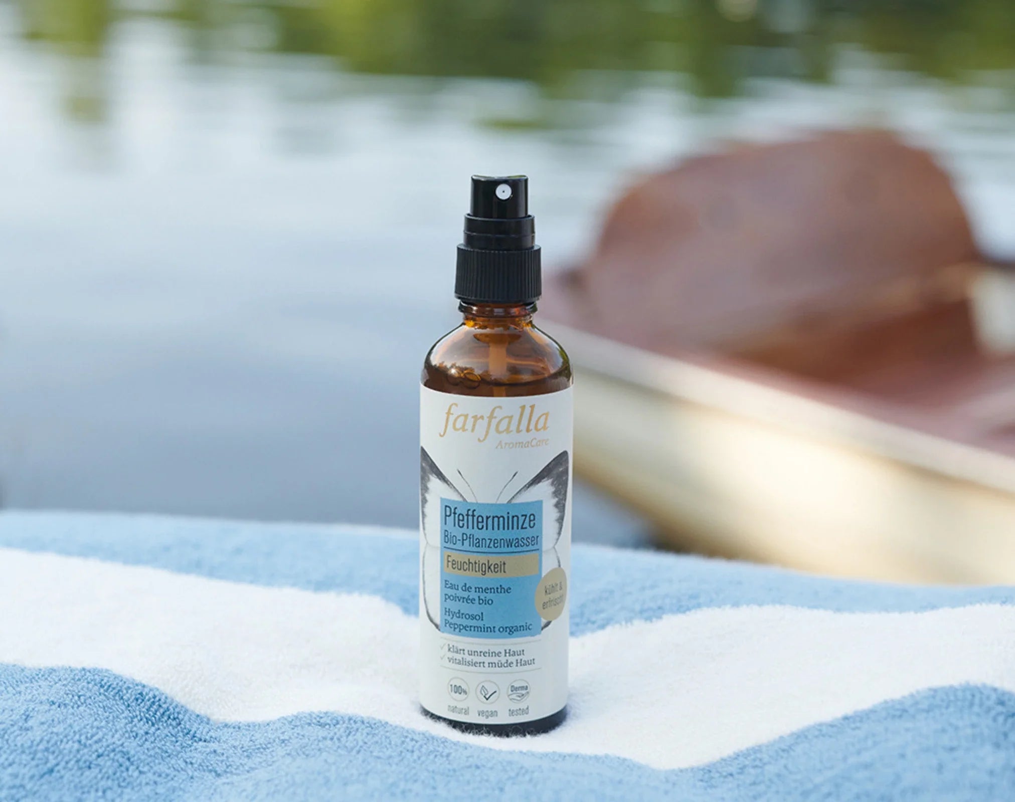 Braunes Sprayfläschchen mit Pfefferminze-Öl auf blau-weiß gestreiftem Handtuch am See, Galerie Position 1, zertifizierte Naturkosmetik